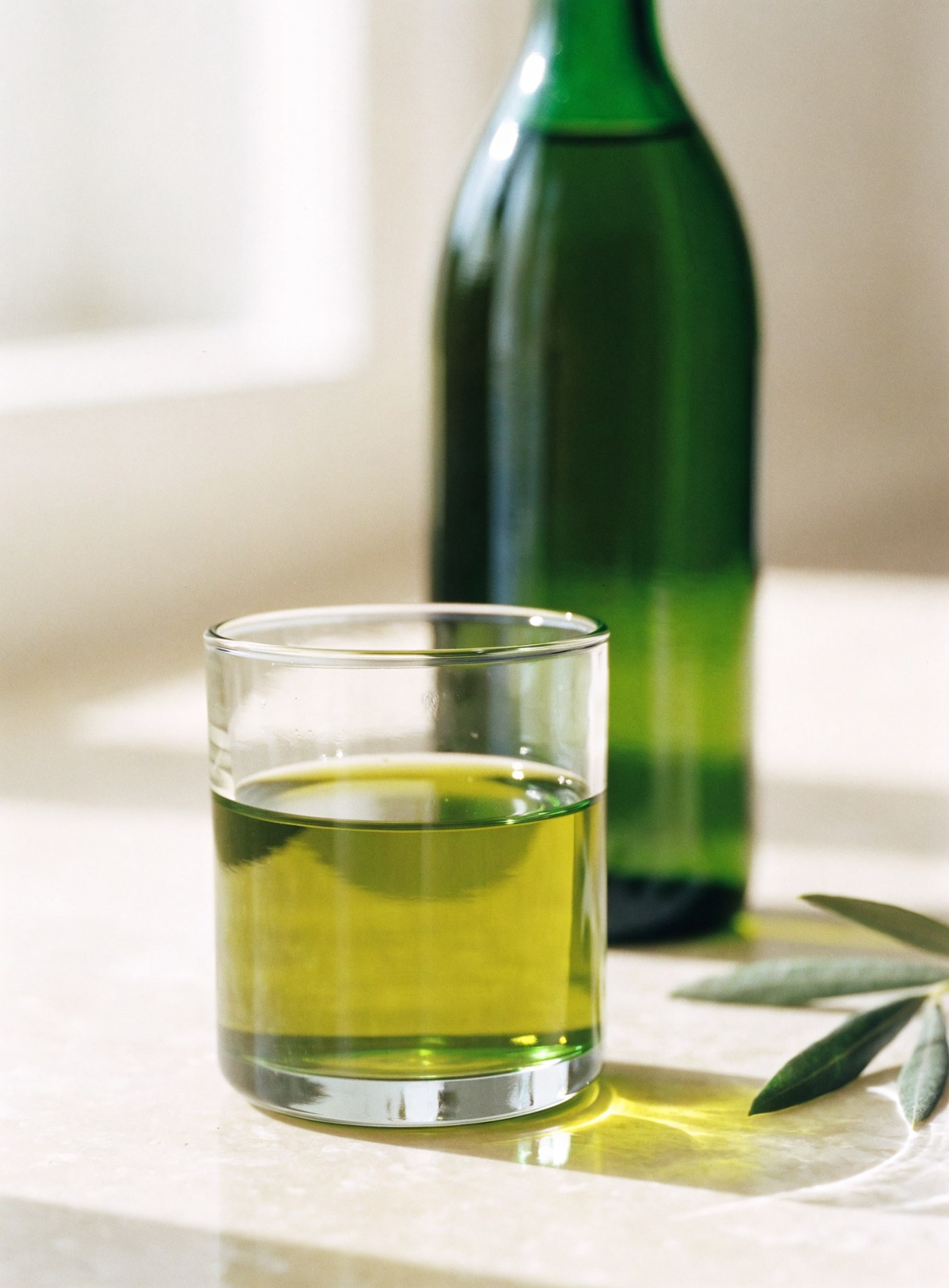 A glass of vivid green olive oil beside a dark bottle and fresh olive leaves