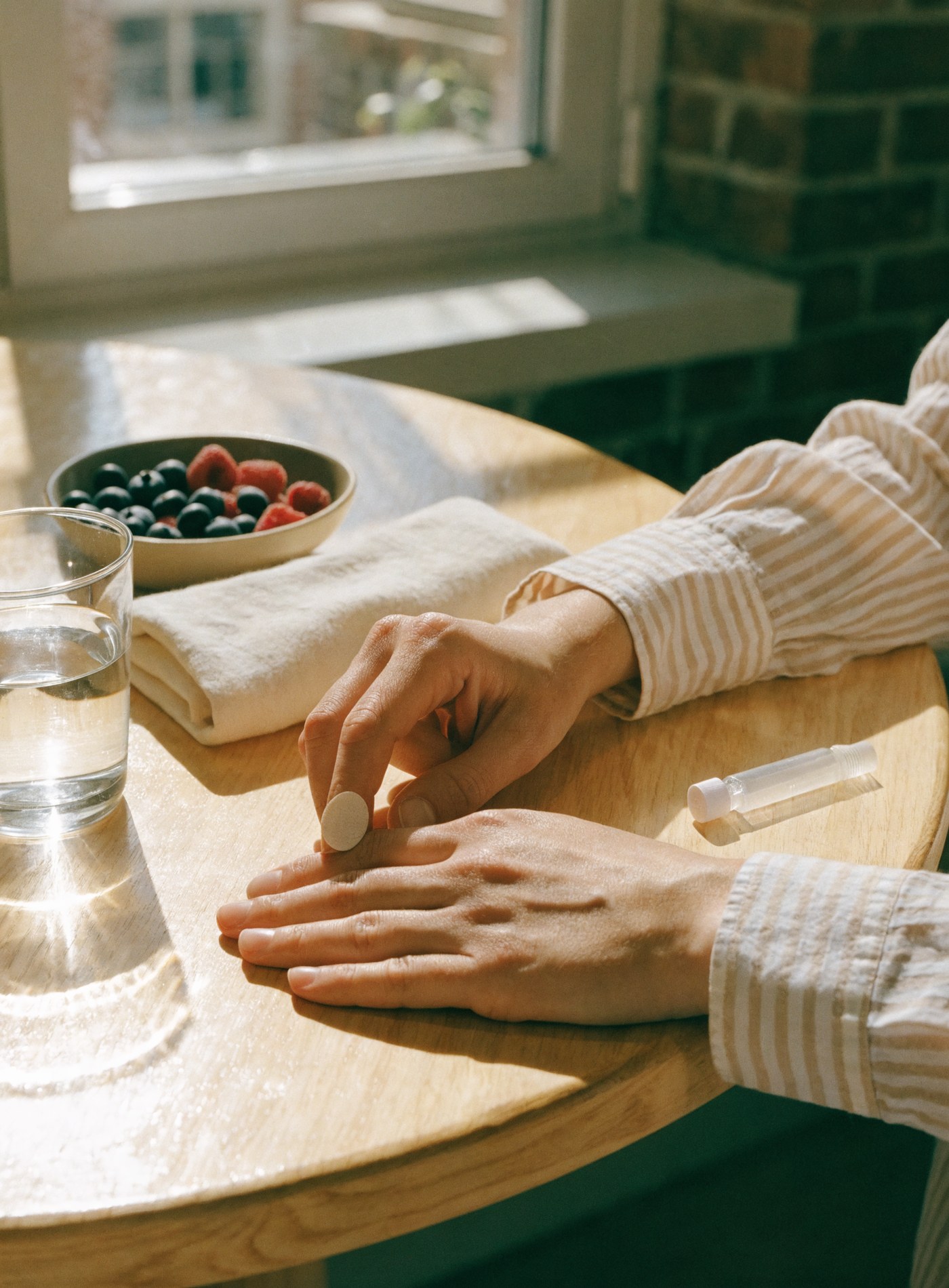 Hands at a sunlit kitchen table, one fingertip with a small plaster, beside a sealed sample tube